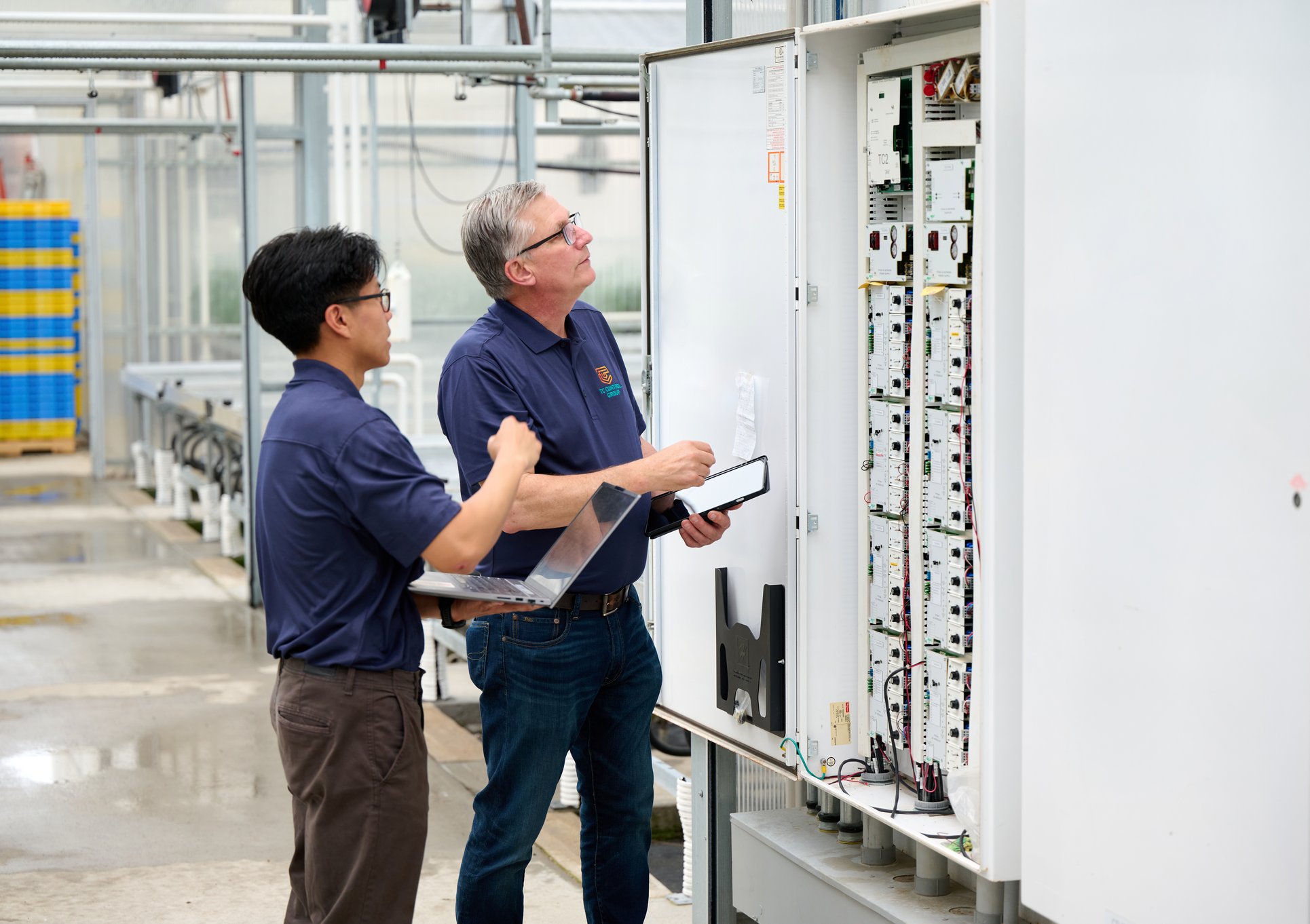 Two TC Controls technicians inspecting an open Argus control panel inside the Nourse Farms greenhouse
