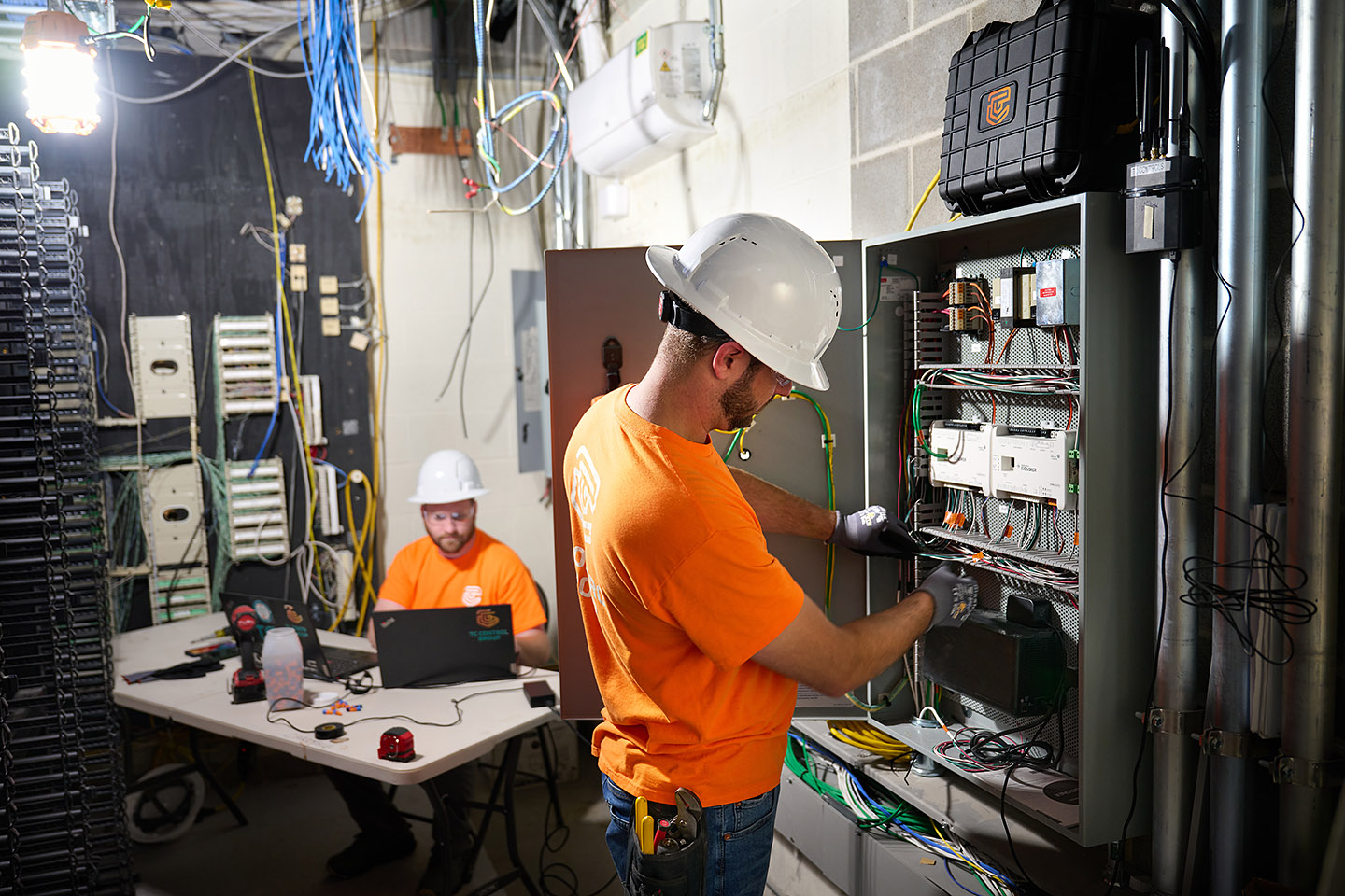 TC Controls technicians terminating field wiring on a mechanical room control panel while programming at a laptop